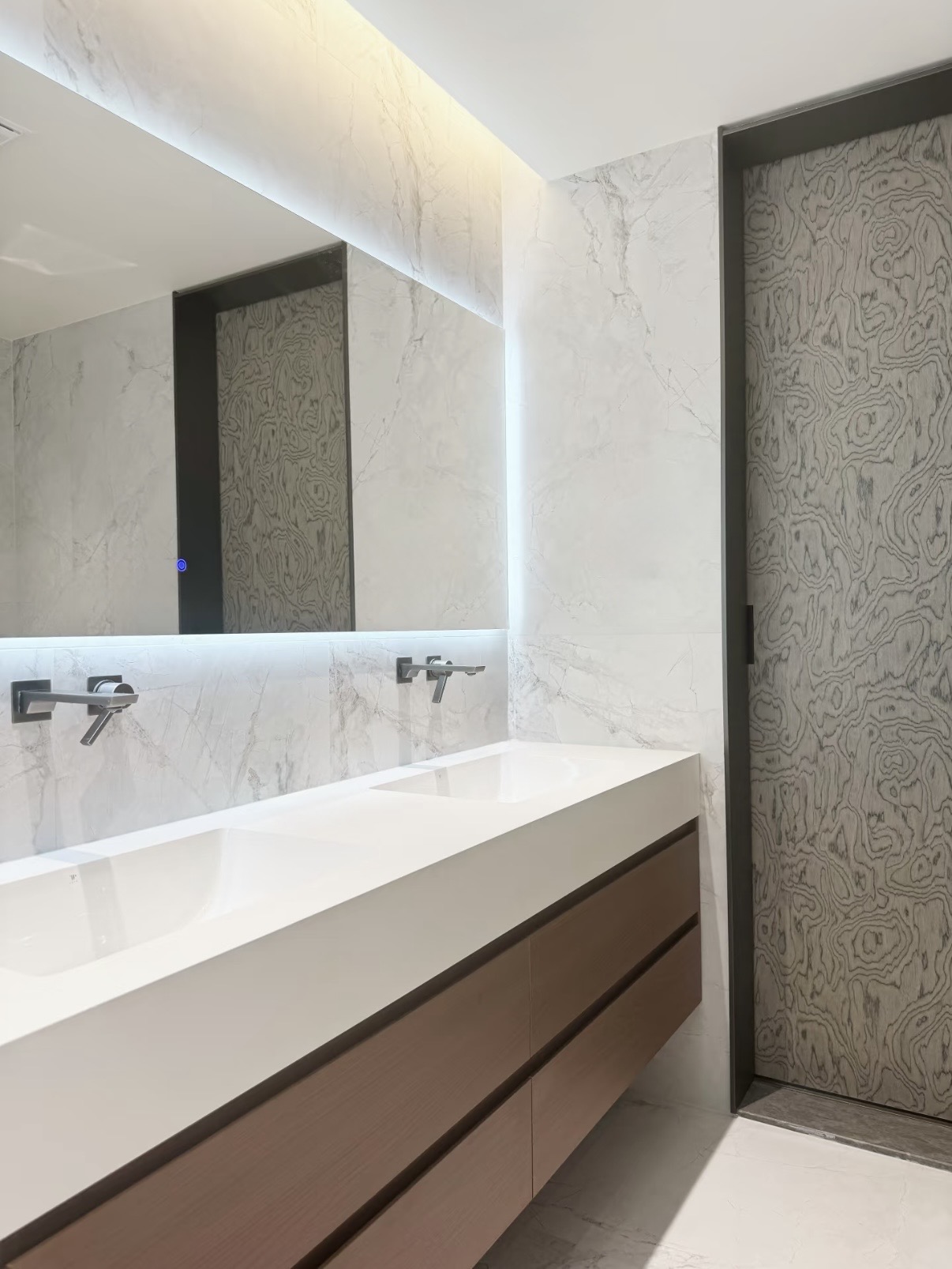 Lave double basin with sintered stone surface, dual wall-mounted taps and LED backlit mirror against stone-clad walls